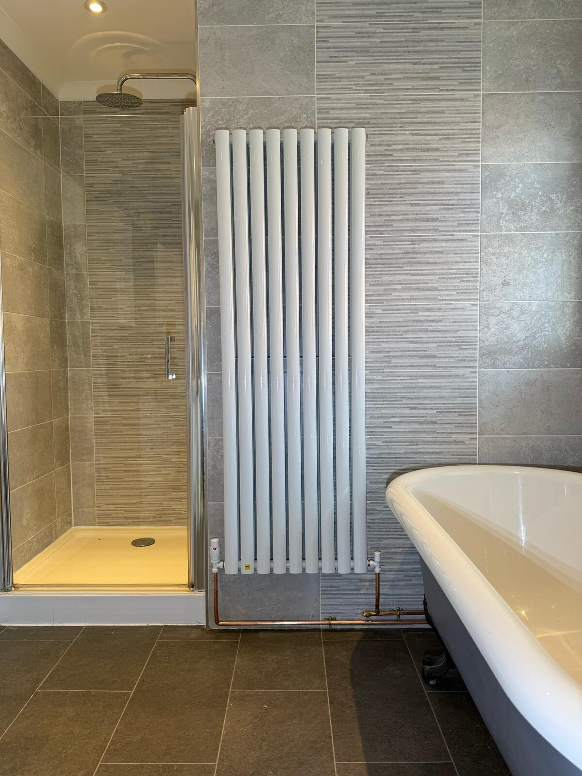 Tall vertical white column radiator installed against grey tiled bathroom wall — bathroom heating upgrade by On-Tap Plumbing & Heating